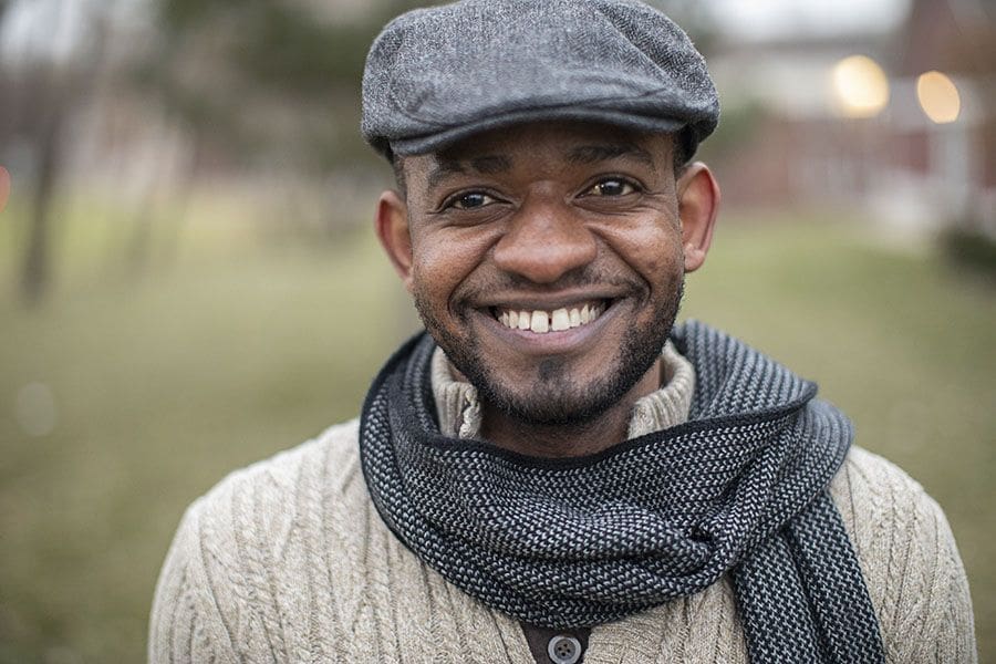 A man with brown skin and a warm smile in a gray scarf and hat