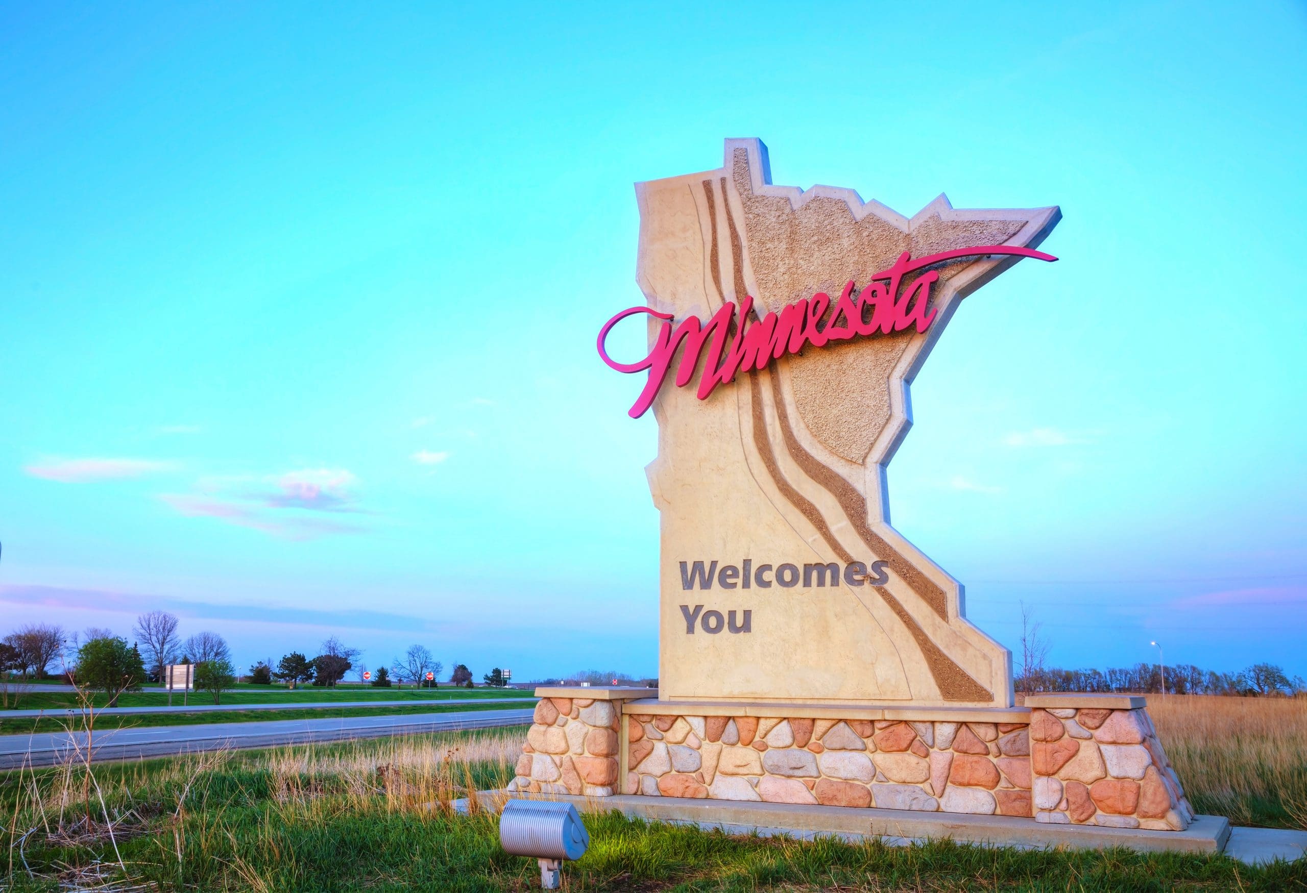 Image of a highway road sign in the shape of the state of Minnesota. It says Minnesota Welcomes You.