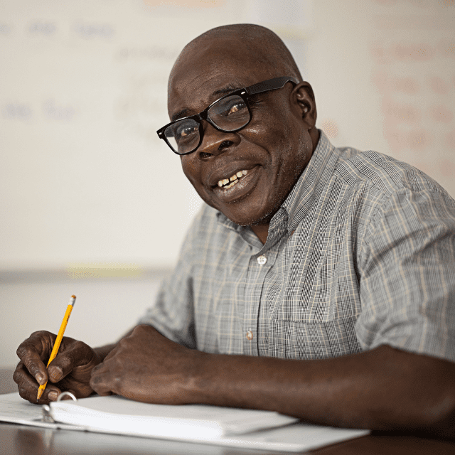 A man writing in class looks up and smiles at the camera.