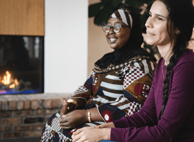 Two women sit together in front of a fireplace, looking ahead and smiling.