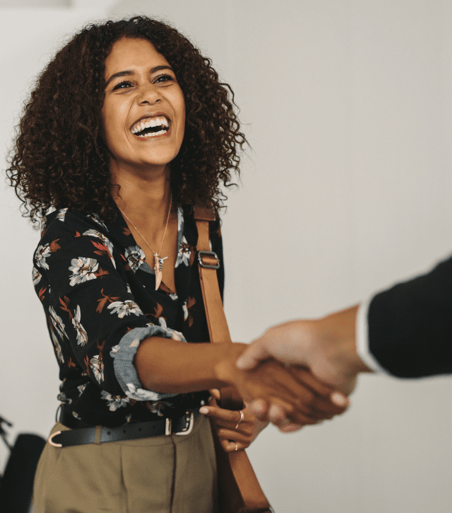 A woman with curly hair smiles and shakes someone's hand in a professional setting.